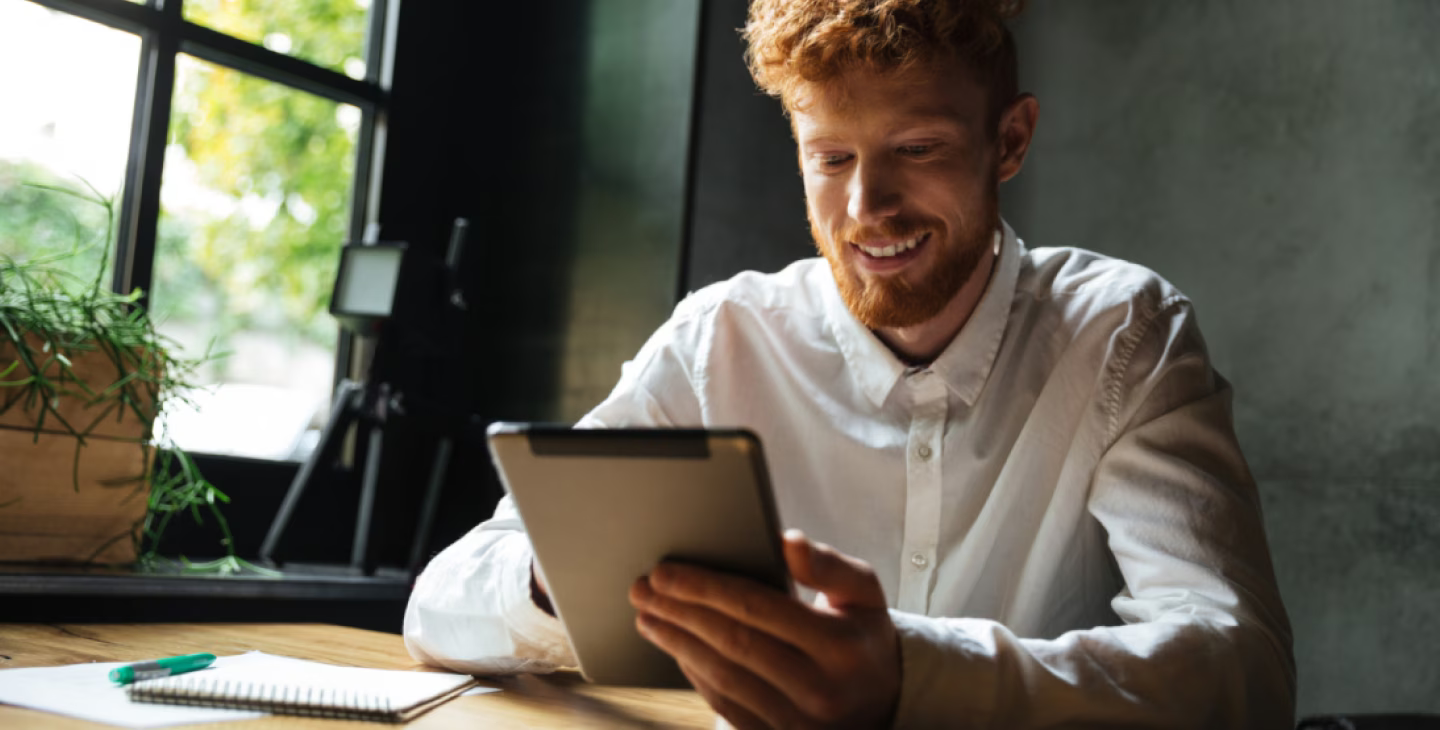 Homme jeune avec cheveux roux, souriant, utilisant une tablette dans un café lumineux avec un carnet et un stylo sur la table.