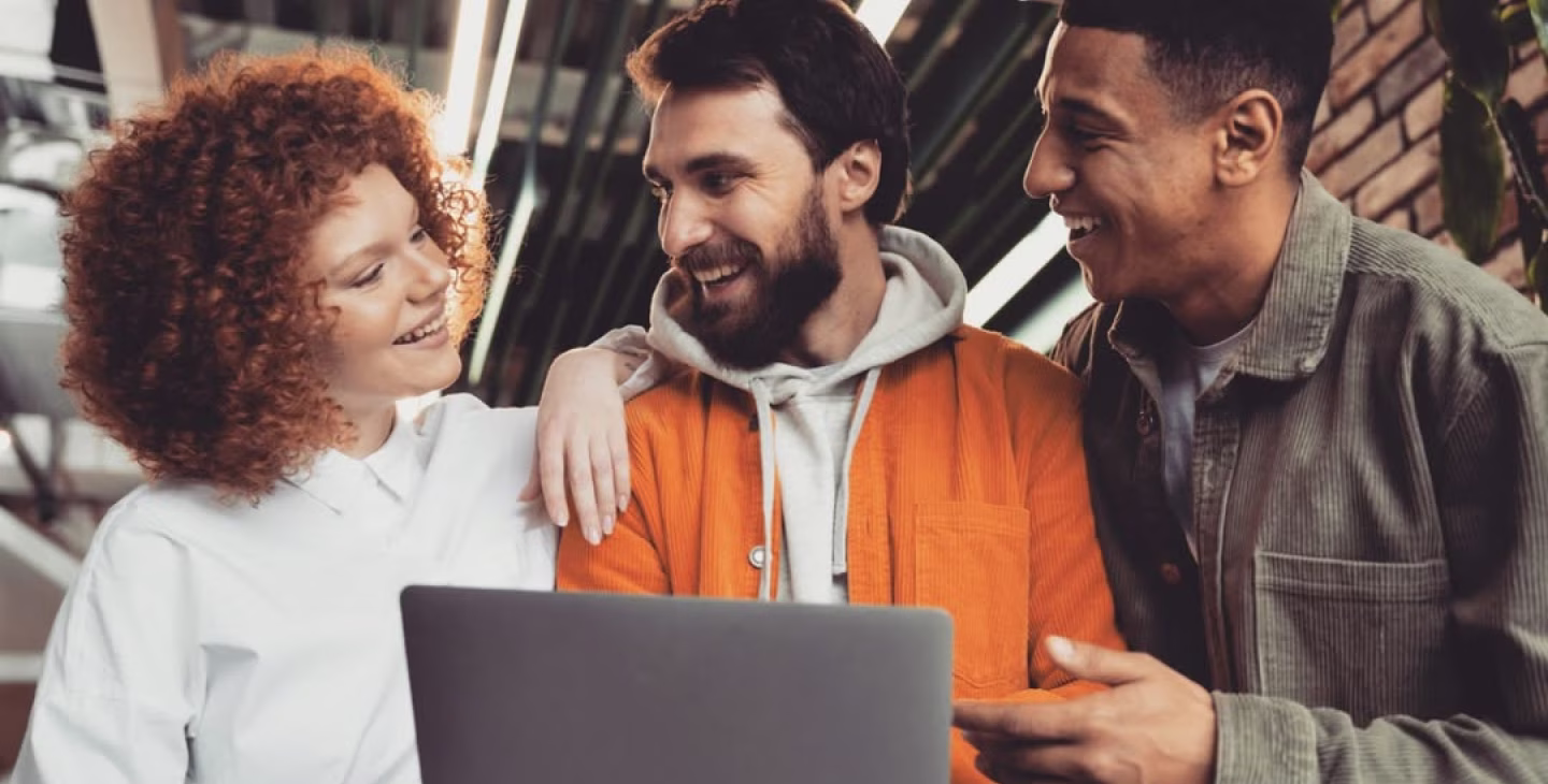 Trois jeunes professionnels souriants discutent et collaborent autour d’un ordinateur portable dans un environnement de travail moderne.