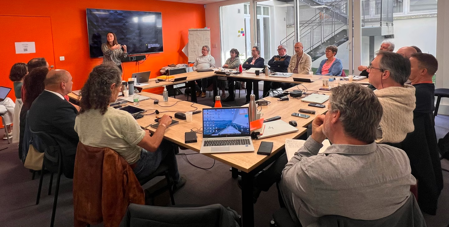 Un groupe de personnes assises autour d'une table de réunion, avec un écran de présentation au fond. Une personne est en train de donner une présentation pendant que les autres écoutent attentivement.