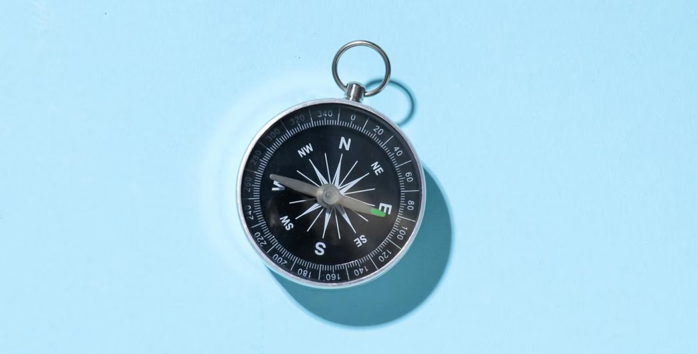 Boussole ancienne métallique avec cadran noir et aiguilles pointant vers l’est sur fond bleu clair