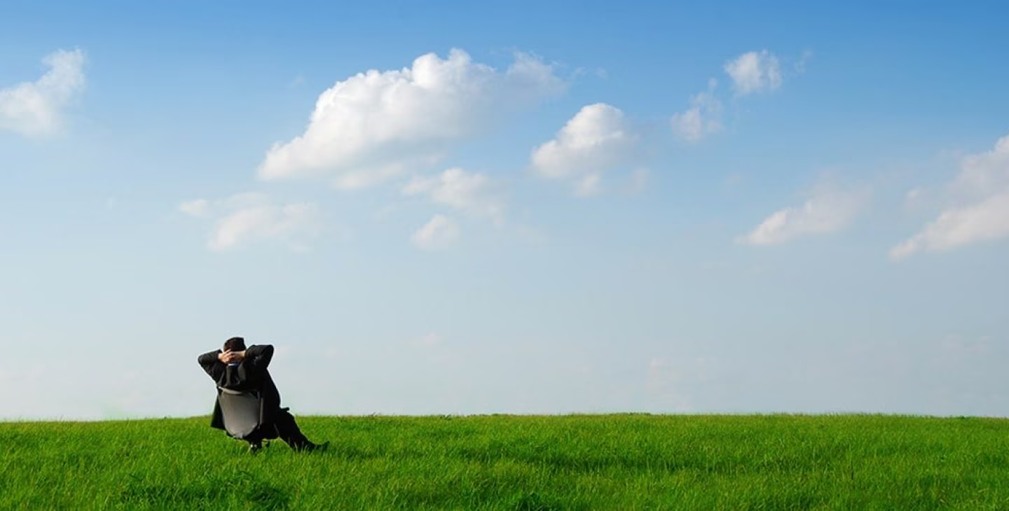 Homme en costume assis dans un fauteuil au milieu d’une prairie verte sous un ciel bleu avec quelques nuages